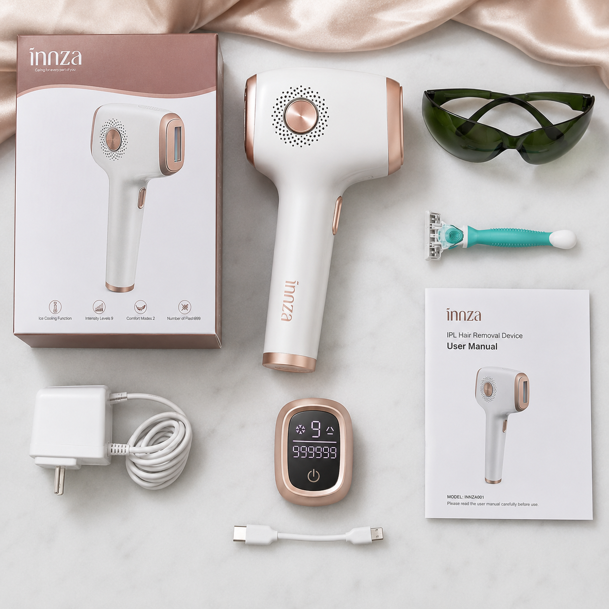 An innza hair removal device set arranged on a light marble surface with satin fabric. The set includes the white and rose gold handheld device, its retail box, a white wall charger, a USB cable, a small digital control pad, the user manual, a teal and white razor, and dark green protective glasses.